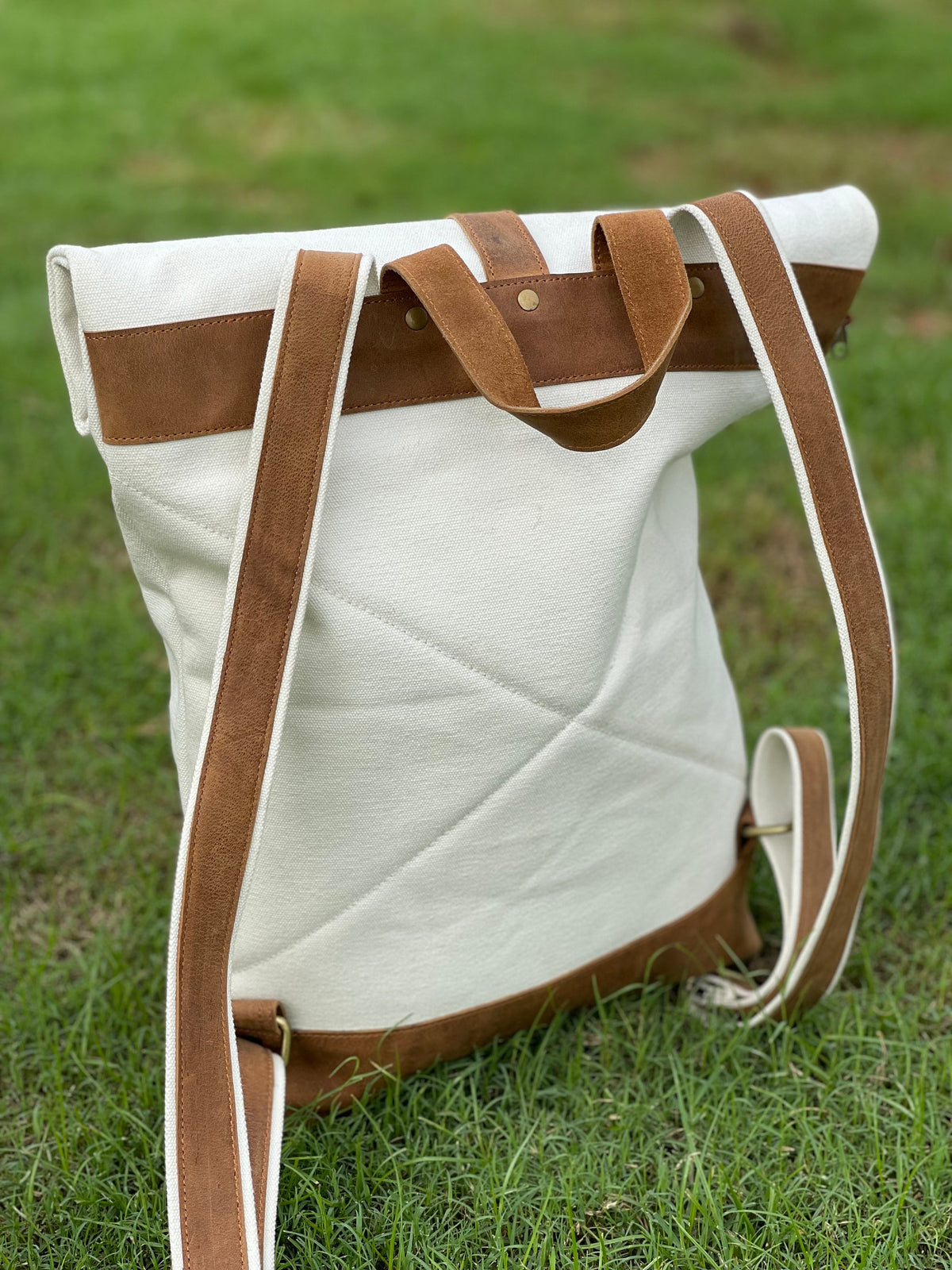 May include: A white canvas backpack with brown leather straps and trim. The backpack has a flap closure and a zippered pocket on the front.