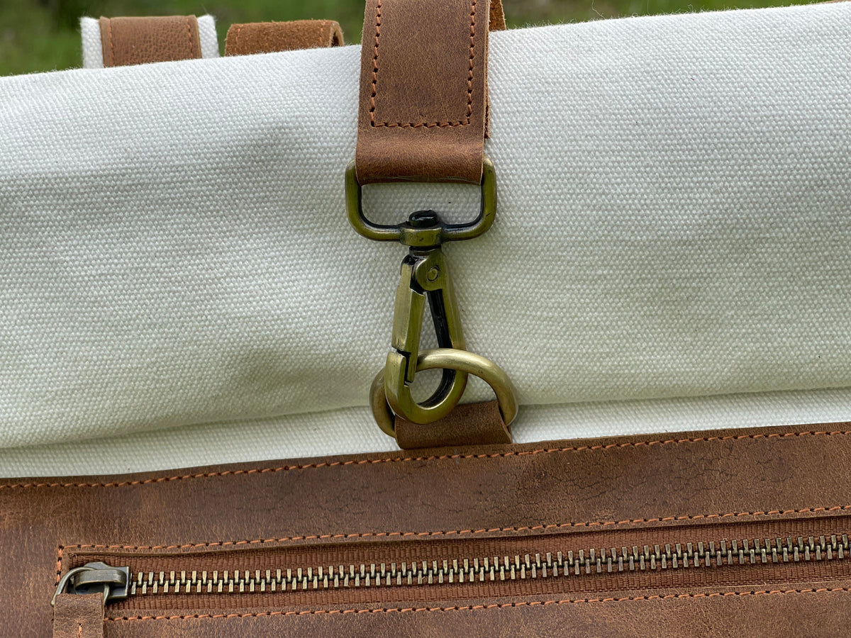 May include: Close-up of a white canvas backpack with a brown leather strap and a brass clasp. The strap is attached to the bag with a zipper.