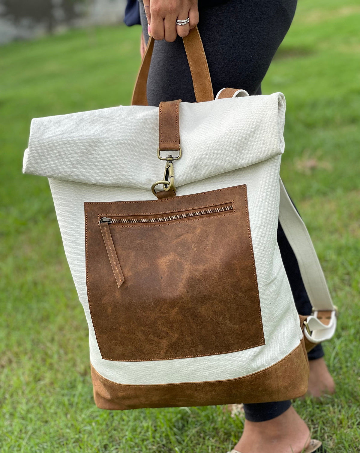 May include: A white canvas backpack with a brown leather flap and a brown leather pocket. The backpack has a roll-top closure and a zippered pocket on the front.