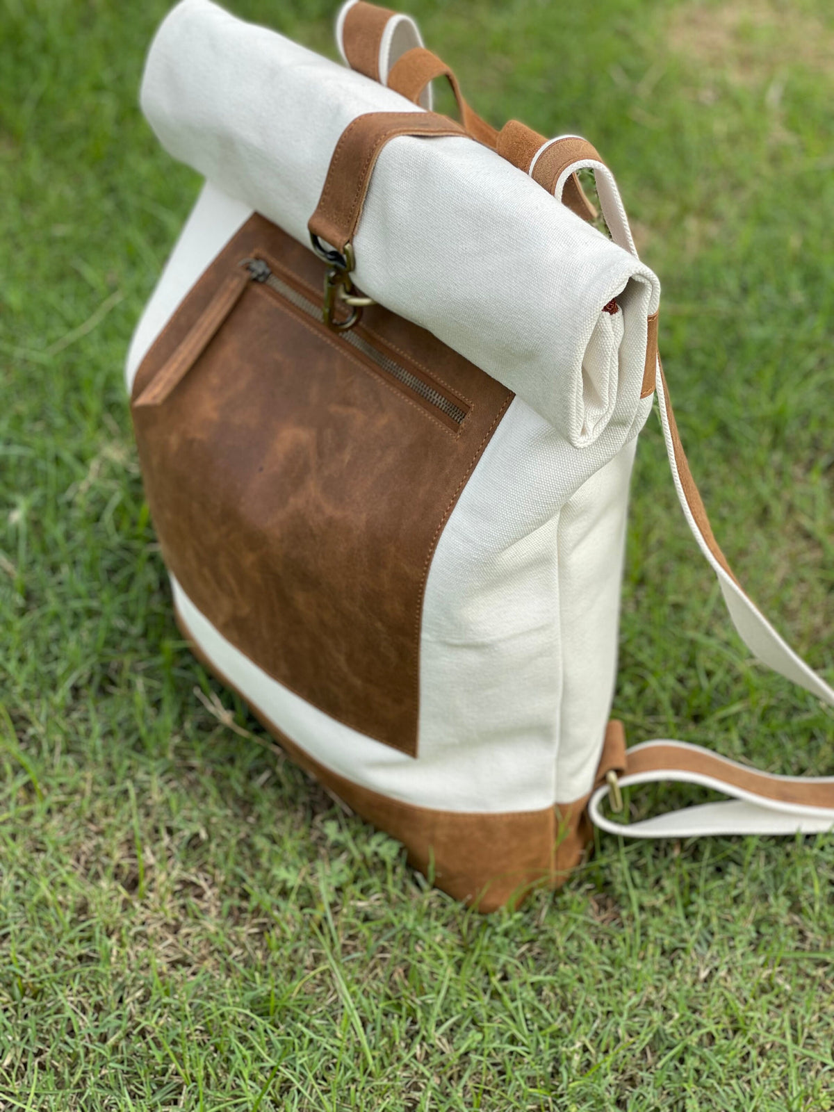 May include: A white canvas backpack with a brown leather flap and a zippered pocket. The backpack has a roll-top closure and adjustable straps.