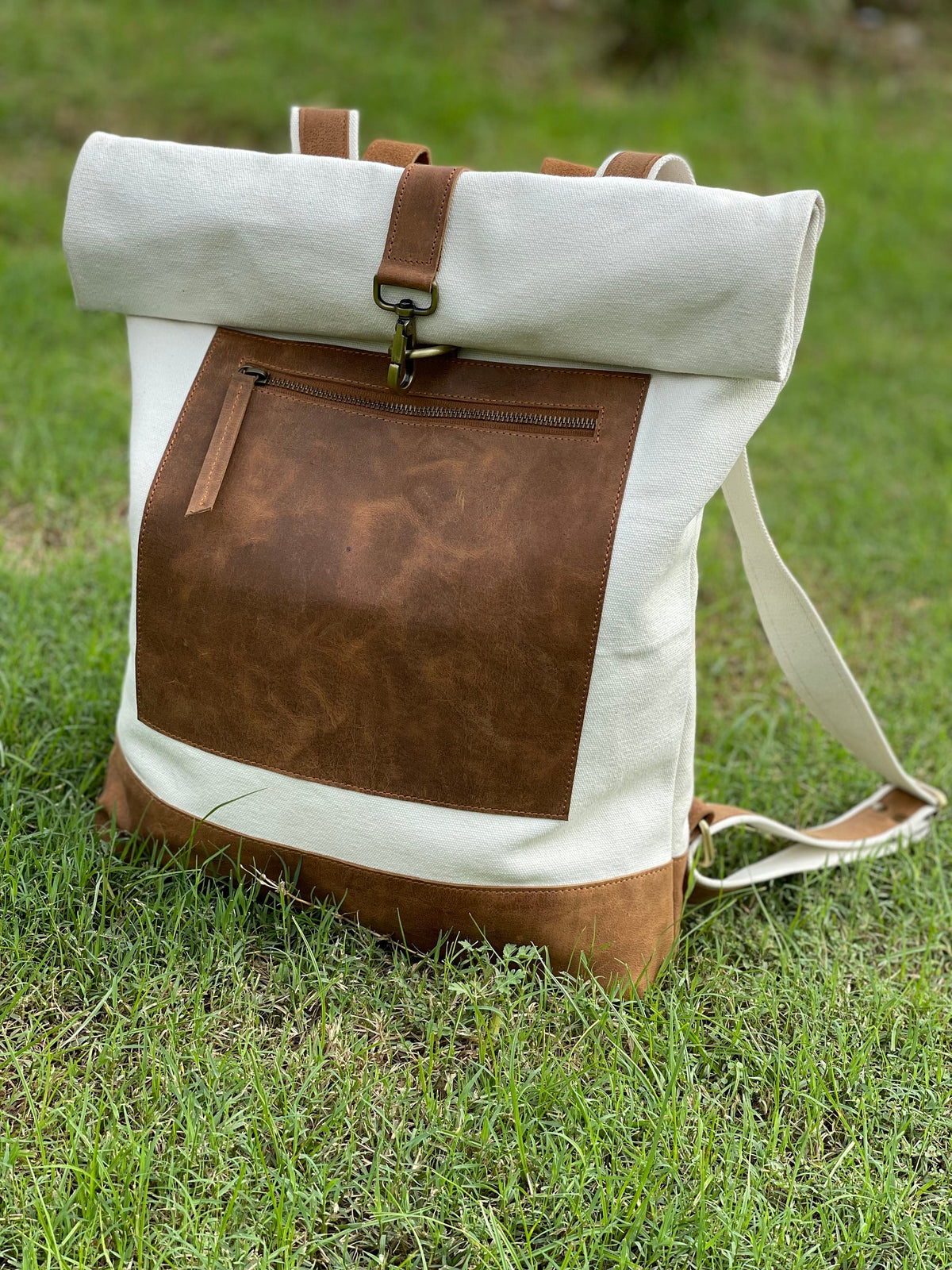 May include: A white canvas backpack with a brown leather flap and a brown leather pocket. The backpack has a roll-top closure and adjustable straps.