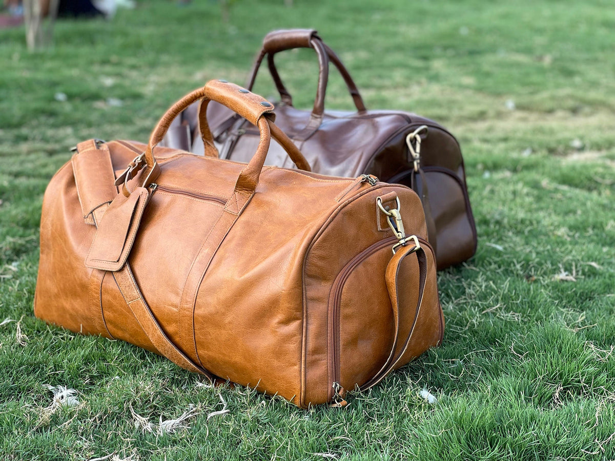 May include: Two leather duffel bags, one brown and one tan, are sitting on a green grassy surface. Both bags have a top handle and a shoulder strap.