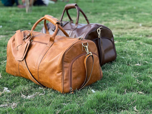 May include: Two leather duffel bags, one brown and one tan, are sitting on a green grassy surface.