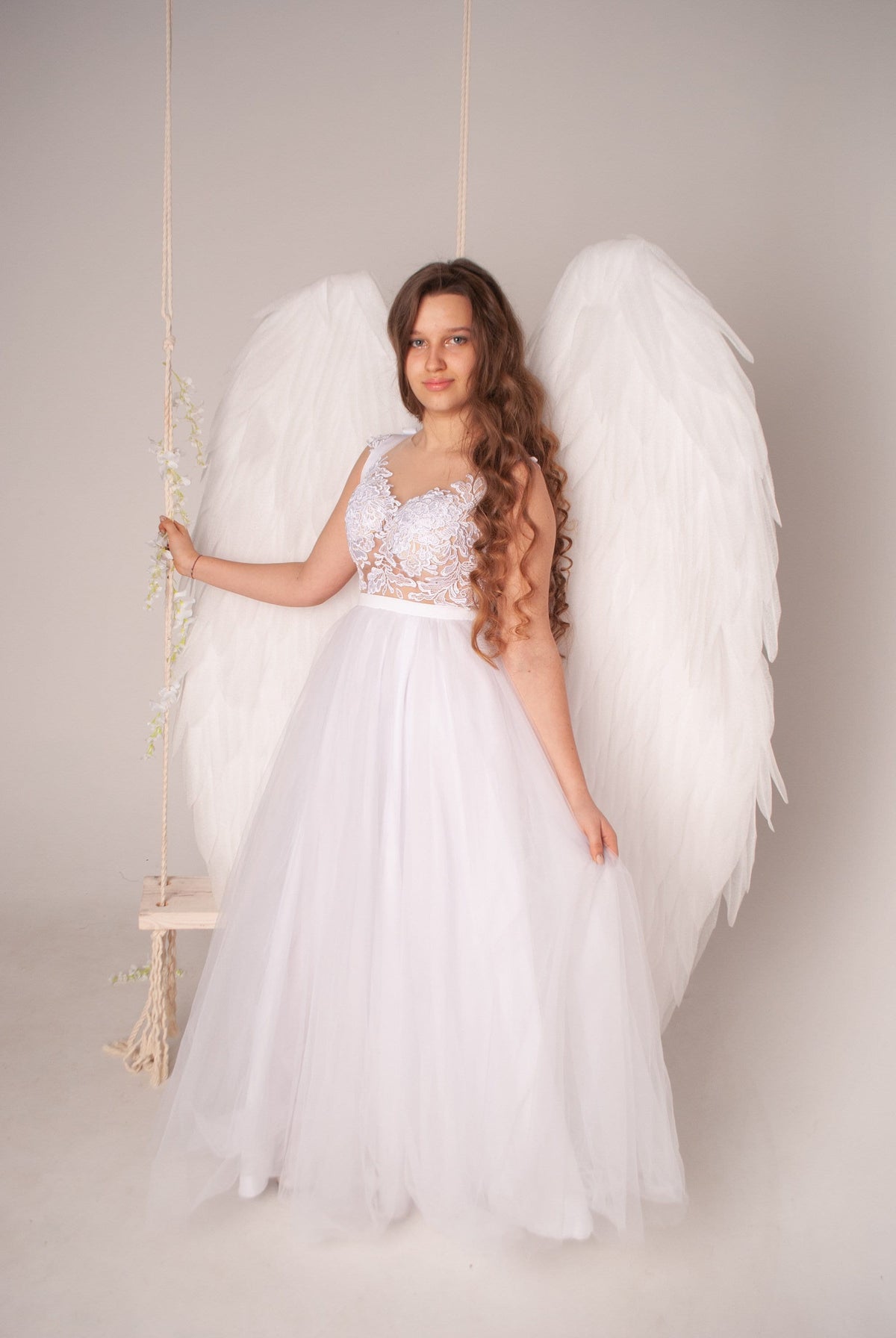 May include: A white tulle dress with a lace bodice and large white angel wings. A swing with floral accents hangs in the background. The dress is floor-length and the wings are large and feathery.