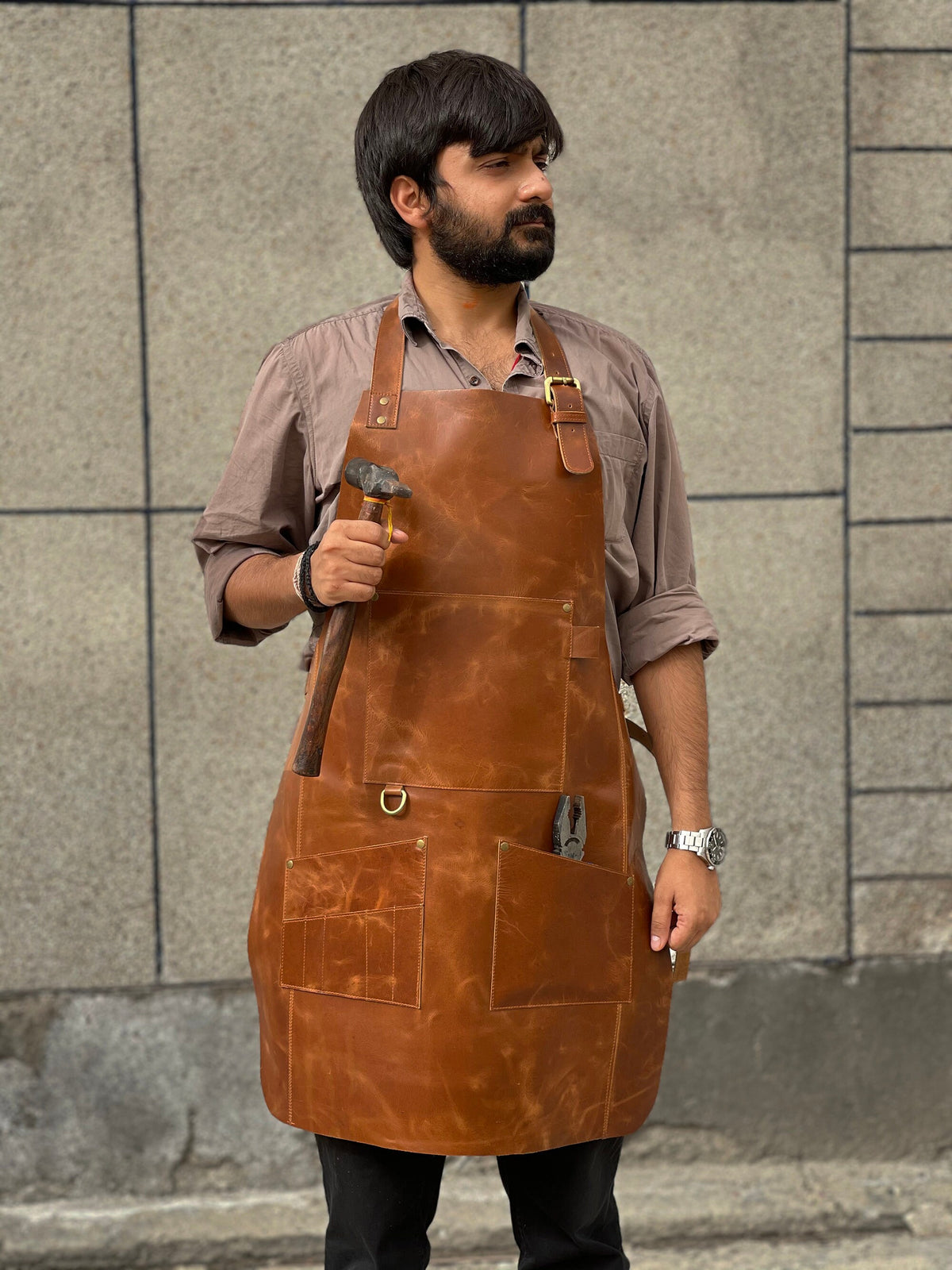 May include: A brown leather apron with multiple pockets and a hammer in the left pocket. The apron has a brass buckle and a brass D-ring.