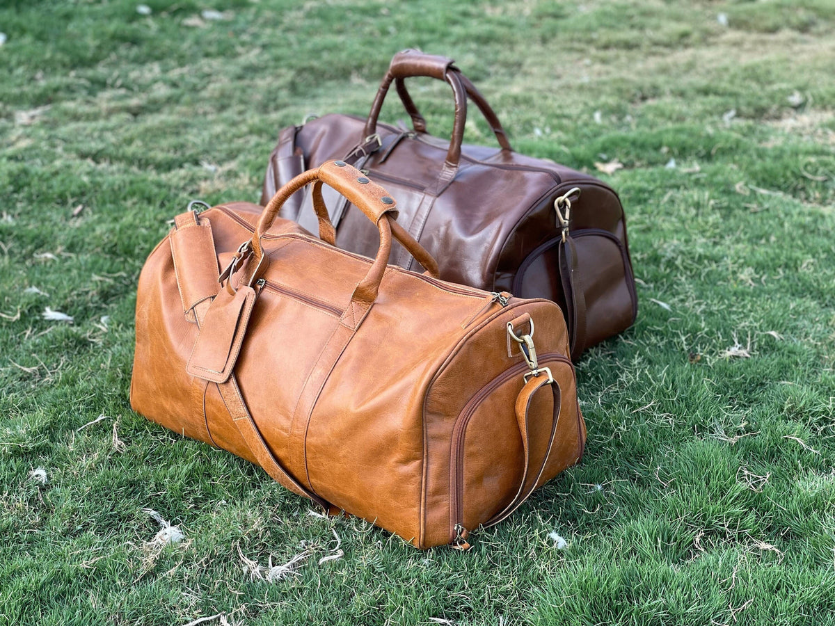 May include: Two leather duffel bags, one brown and one tan, are sitting on a bed of green grass. Both bags have a top handle and a shoulder strap.