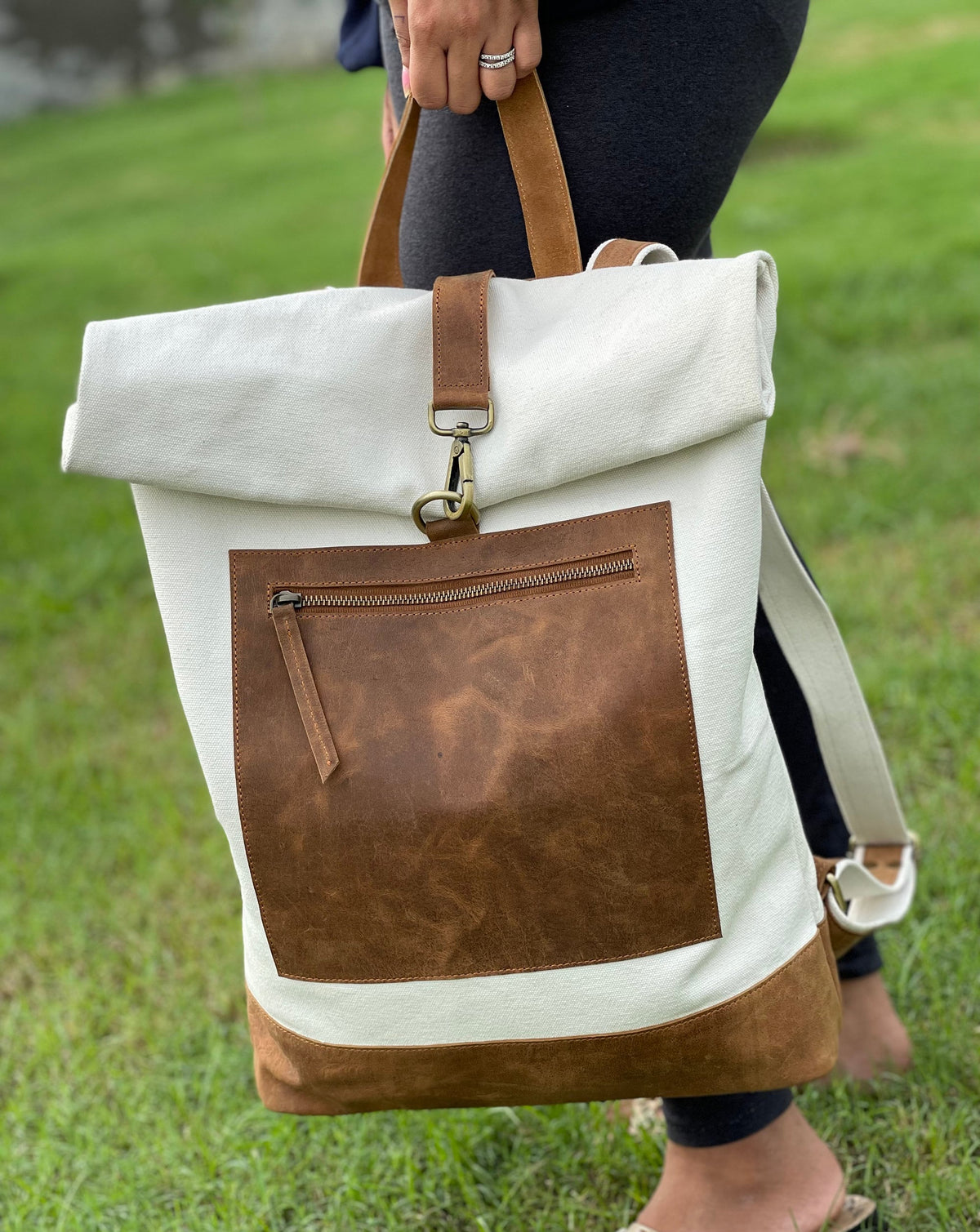 May include: A white canvas backpack with a brown leather flap and a brown leather bottom. The backpack has a zippered pocket on the front and a large main compartment.