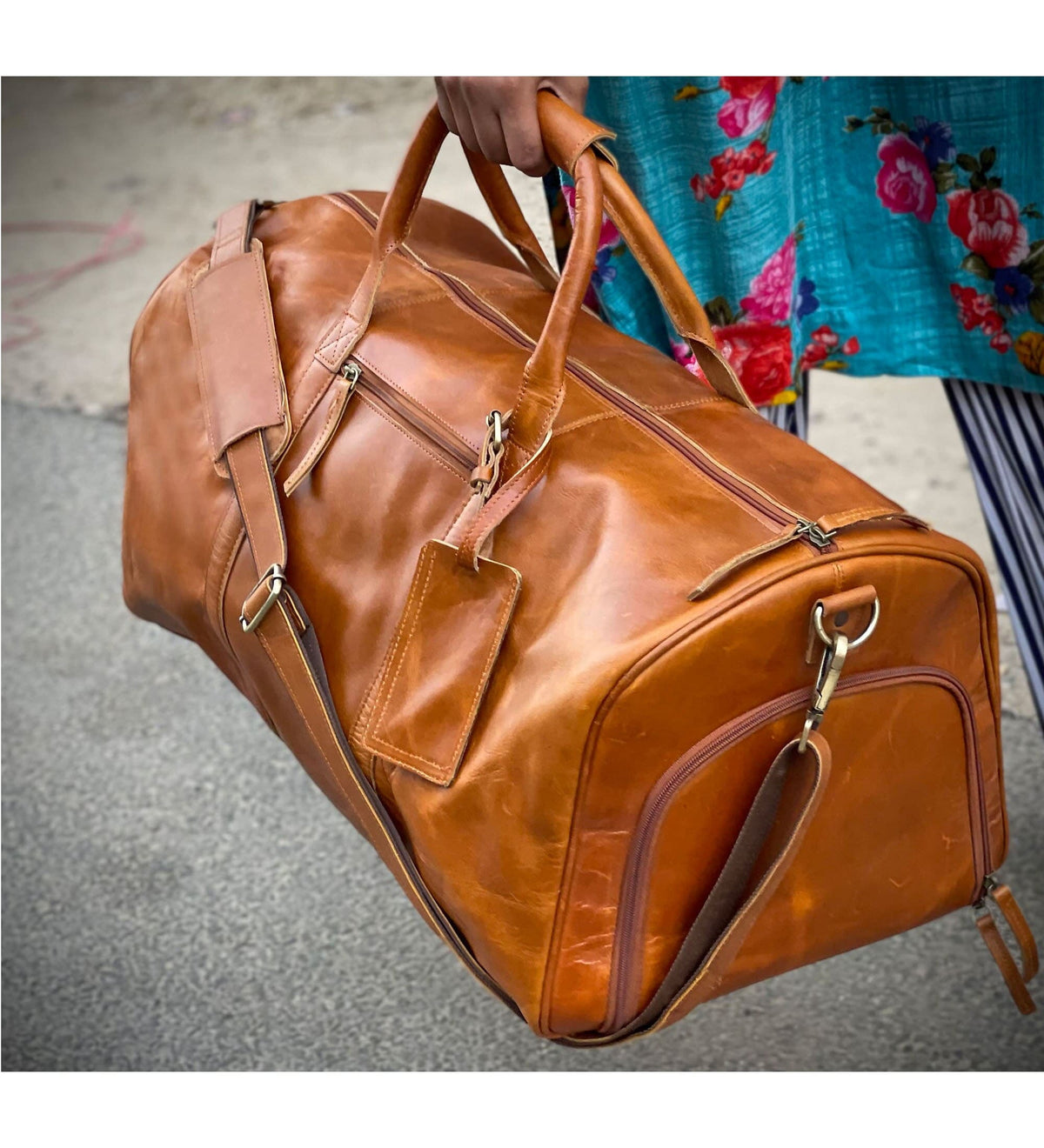 May include: A brown leather duffel bag with a long shoulder strap and two top handles. The bag has a zipper closure and a small pocket on the front.