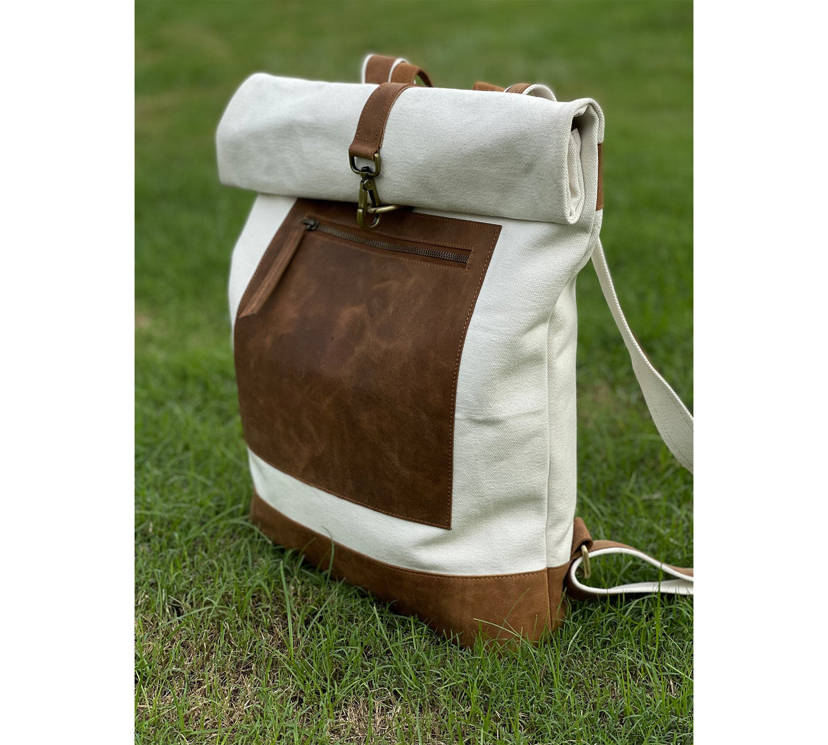 May include: A white canvas backpack with a brown leather flap and a brown leather pocket. The backpack has a roll-top closure and adjustable straps.