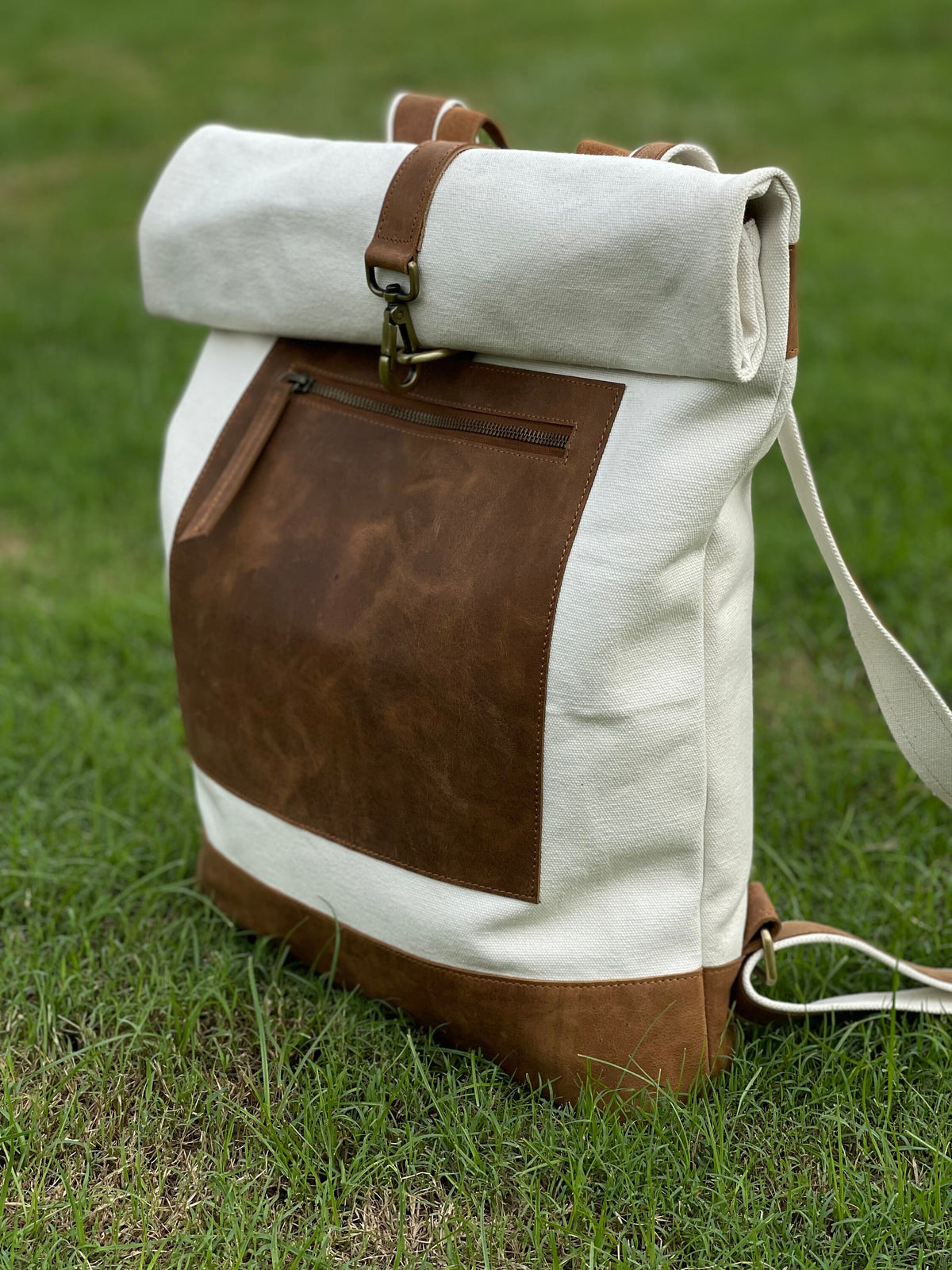 May include: A white canvas backpack with a brown leather flap and a brown leather pocket. The backpack has a roll-top closure and a metal buckle.