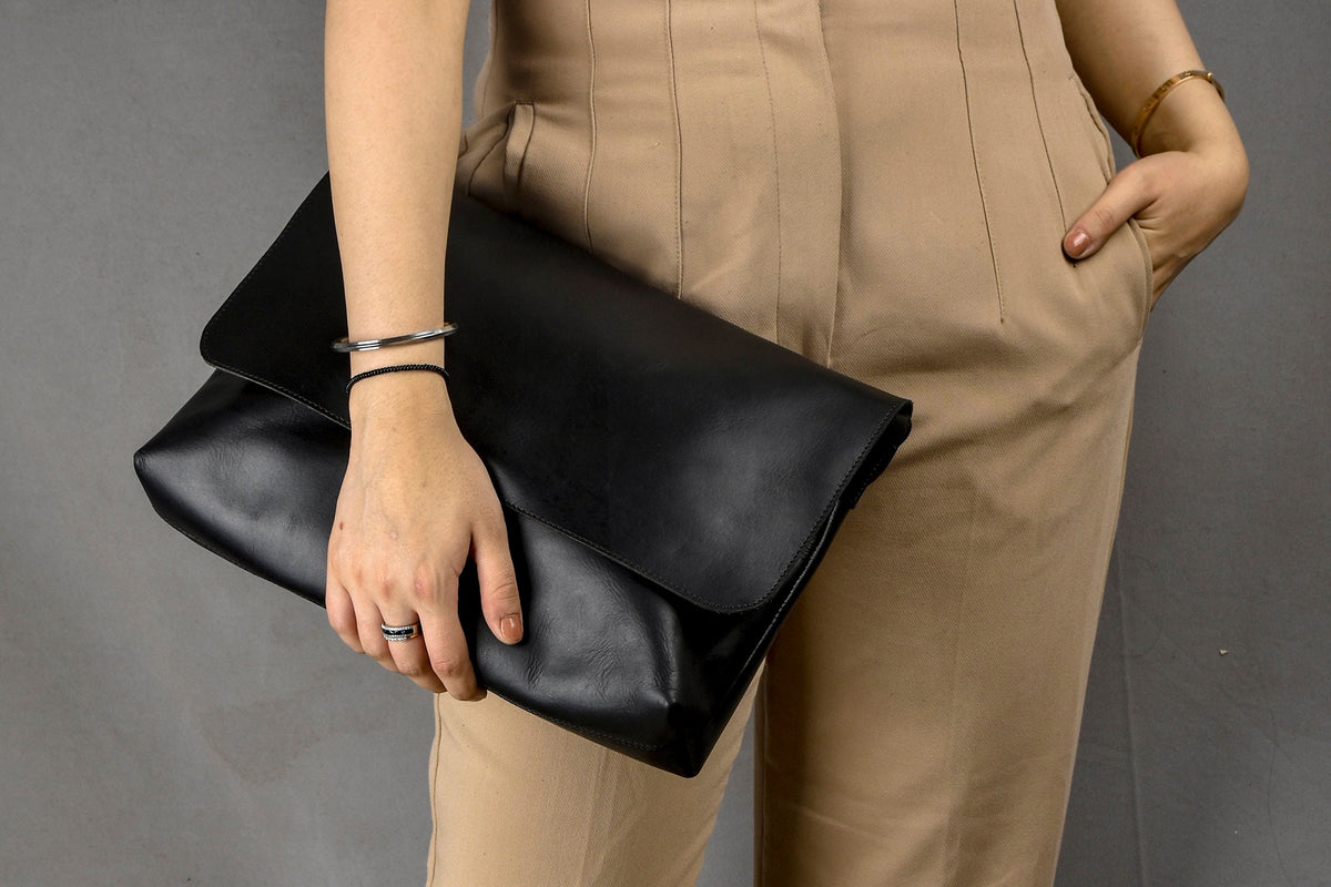 May include: A black leather clutch with a folded flap, held by a person wearing tan pants and a bracelet. The clutch is a solid black color and appears to be made of smooth leather. The background is a solid gray.
