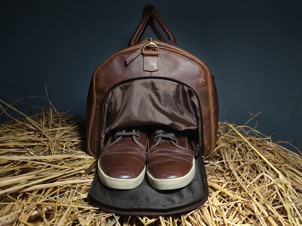 May include: A brown leather duffel bag with a zippered opening, revealing a brown fabric lining. Two brown leather shoes are inside the bag, with the laces tied. The bag is sitting on a bed of straw.