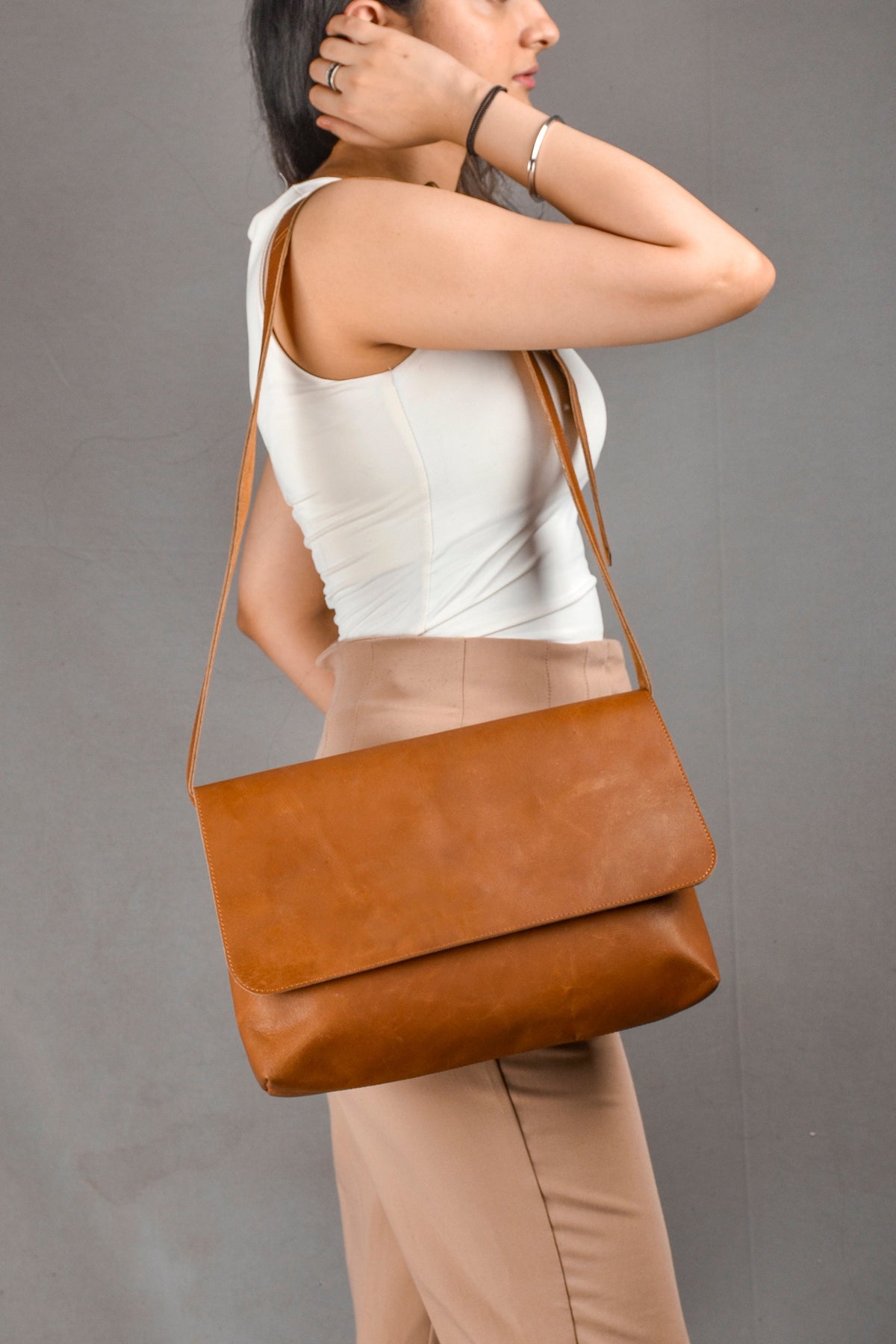 May include: A brown leather crossbody bag with a flap closure. The bag is worn by a person with a white tank top and beige pants.