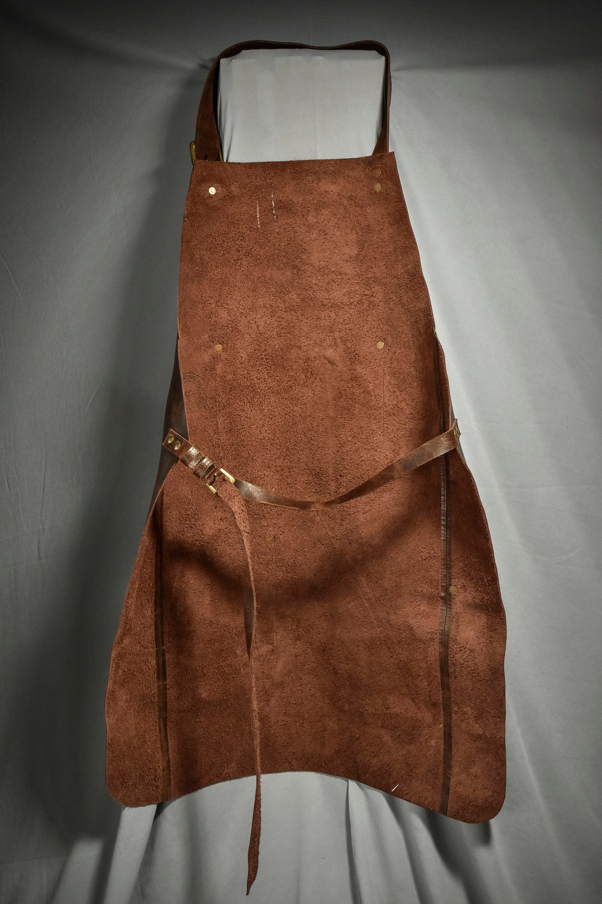 May include: A brown leather apron with adjustable straps and brass buckles. The apron has a suede-like texture and is designed for durability.