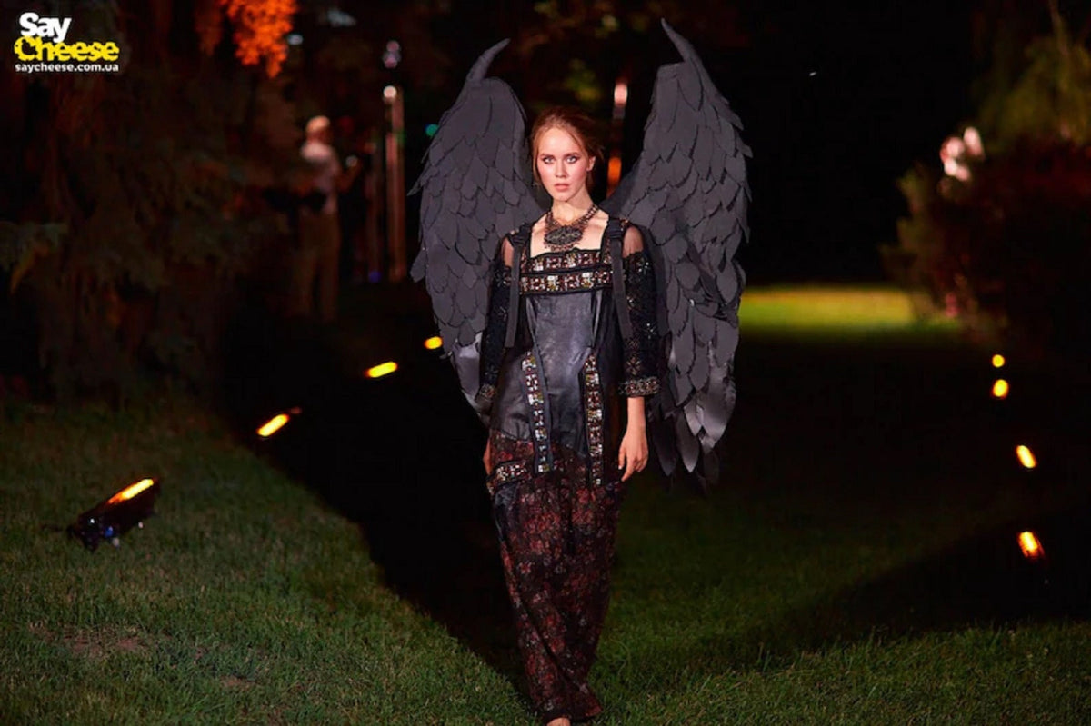 May include: A woman in a dark, ornate costume with large black feathered wings. The dress is black with gold and red accents. The background is a dimly lit outdoor setting with green grass and lights.