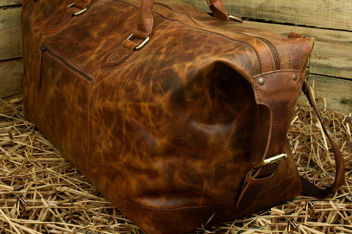 May include: A brown leather duffel bag with brass hardware and a zippered pocket. The bag is sitting on a bed of straw.