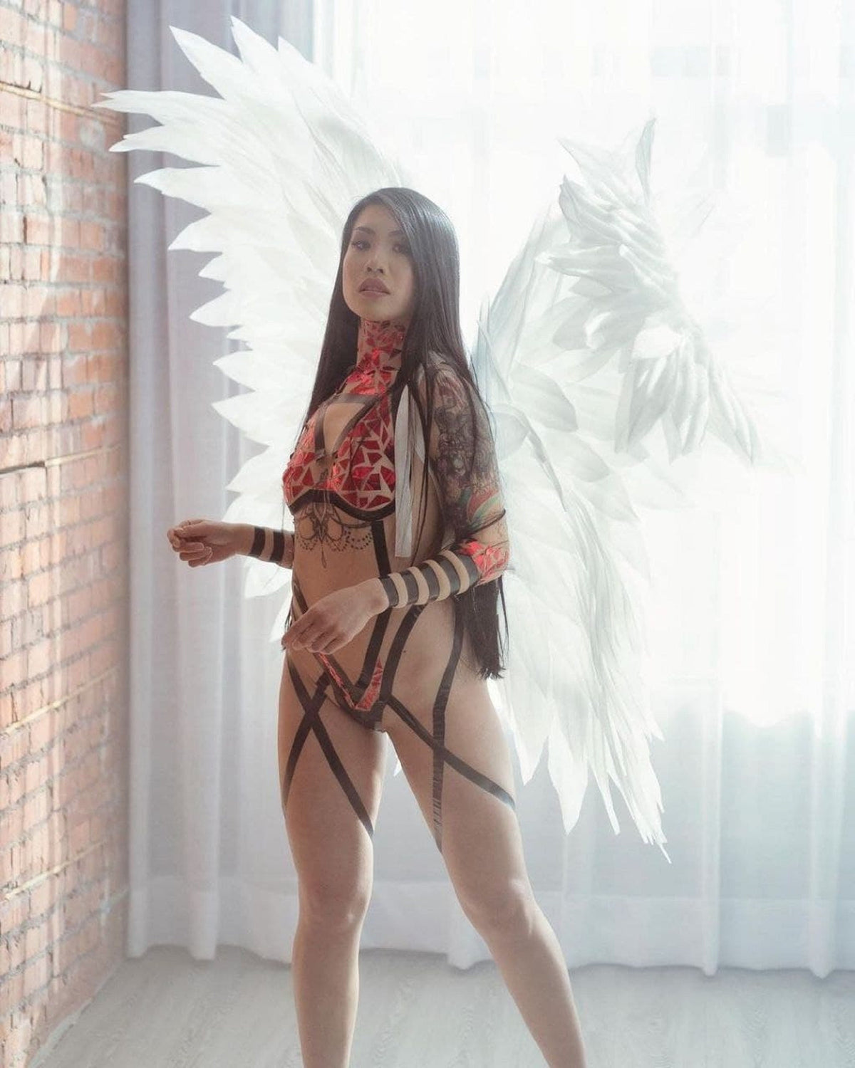 May include: A woman wearing a red and black patterned bodysuit with black straps and a pair of large white angel wings.