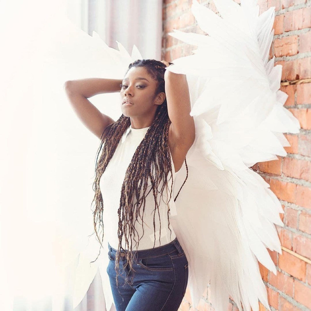 May include: A woman with long brown braids wears a white sleeveless top and blue jeans. She is standing in front of a brick wall with large white angel wings behind her.