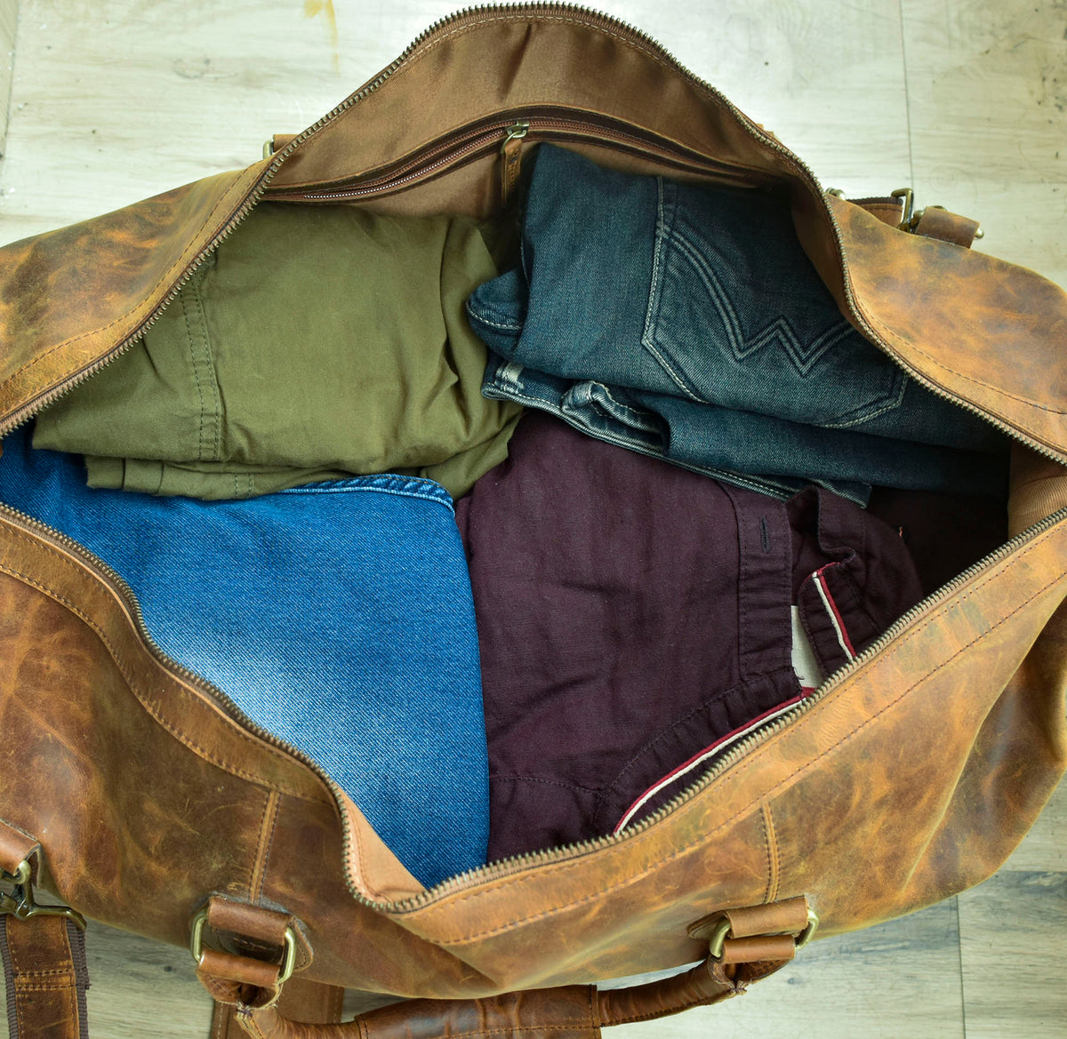 May include: A brown leather duffel bag with a zippered opening. The bag is packed with clothing, including a green jacket, two pairs of blue jeans, and a burgundy shirt.