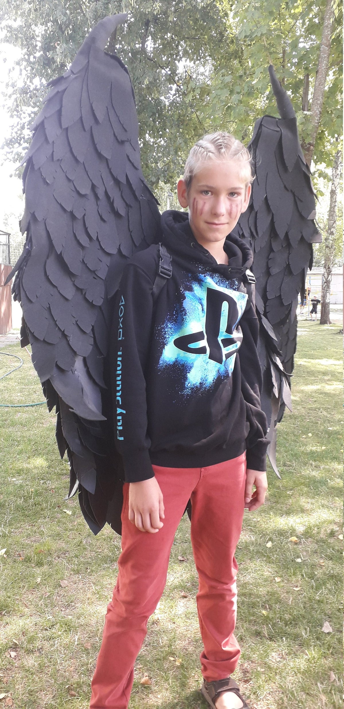 May include: A person wearing a black hoodie with a PlayStation logo and red pants stands outdoors, adorned with large black angel wings. The wings are detailed and add a dramatic flair to the outfit.