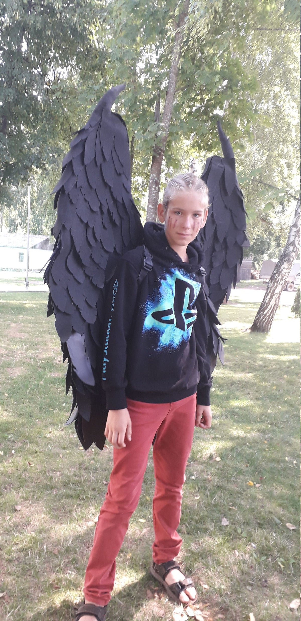 May include: A person wearing large black feathered wings, a black hoodie with a blue PlayStation logo, and red pants. The wings are attached to a backpack. The person is standing on grass.