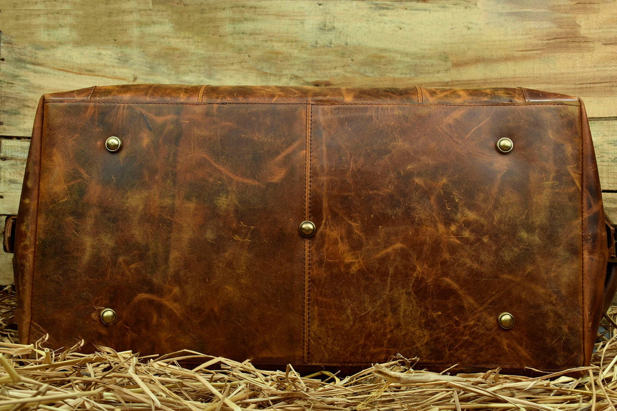 May include: A brown leather duffel bag with brass accents. The bag is sitting on a bed of straw.