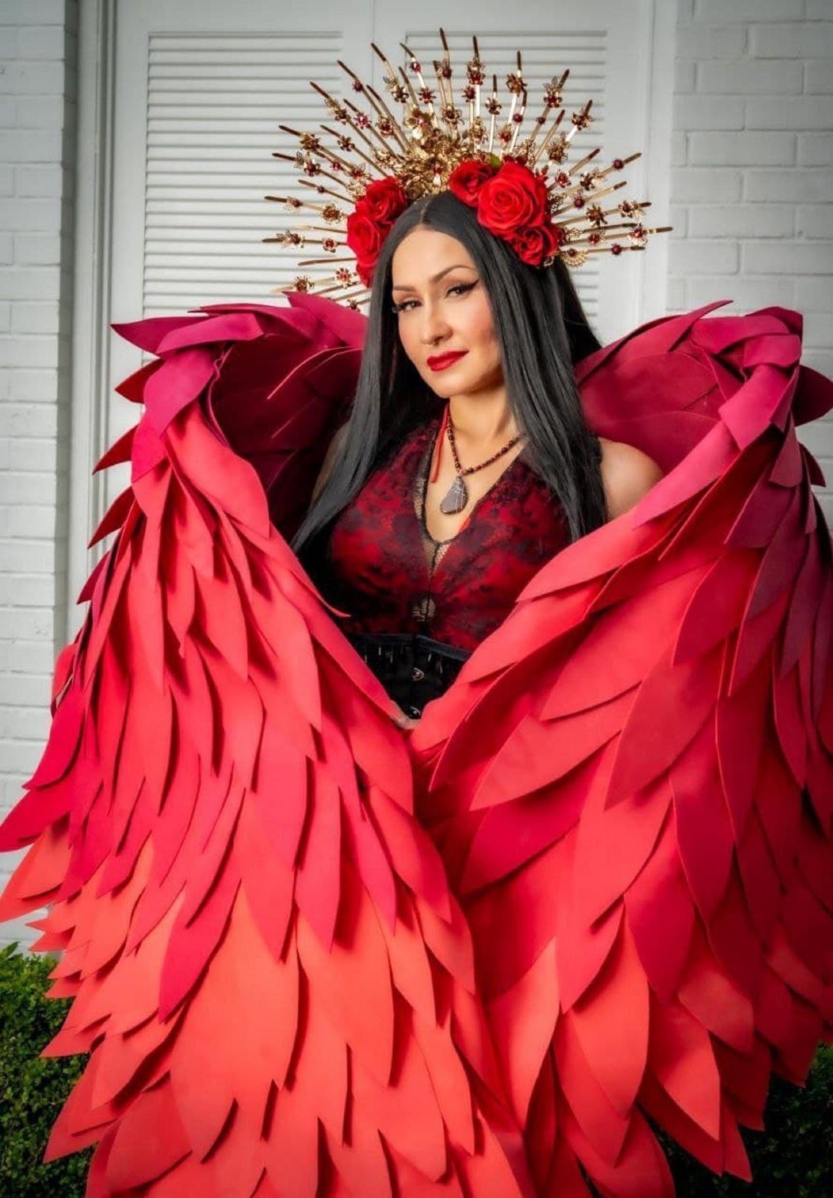 May include: A woman wearing a red, layered, paper angel wing costume with a gold crown adorned with red roses.