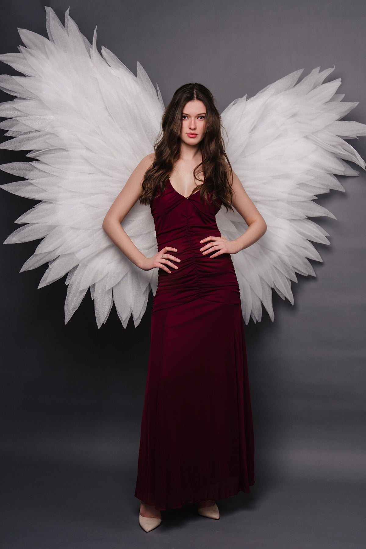 May include: A woman wearing a long, burgundy dress with a pair of white angel wings behind her. She is standing with her hands on her hips and looking directly at the camera.