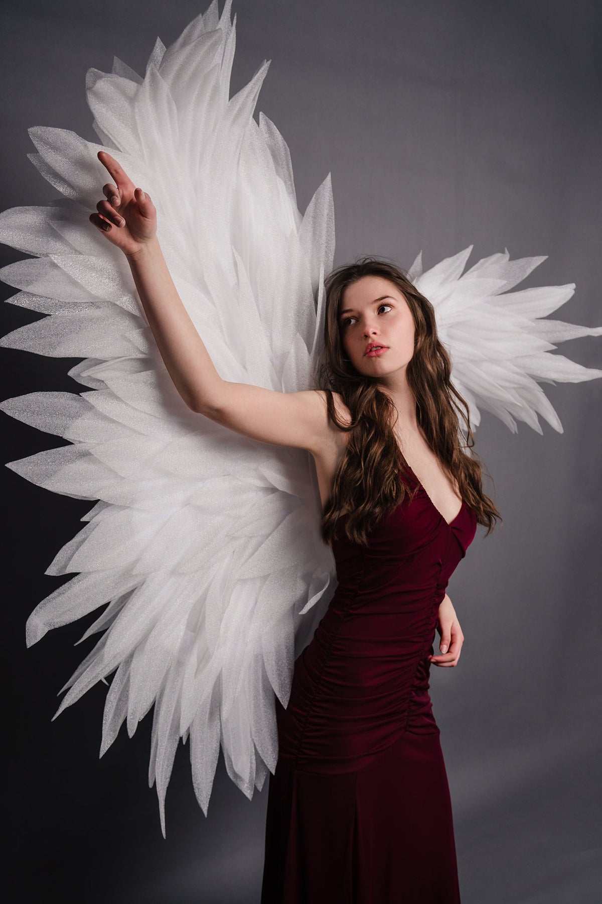 May include: A woman wearing a long, burgundy dress with white angel wings. She is looking up and to the right, with her right arm raised.