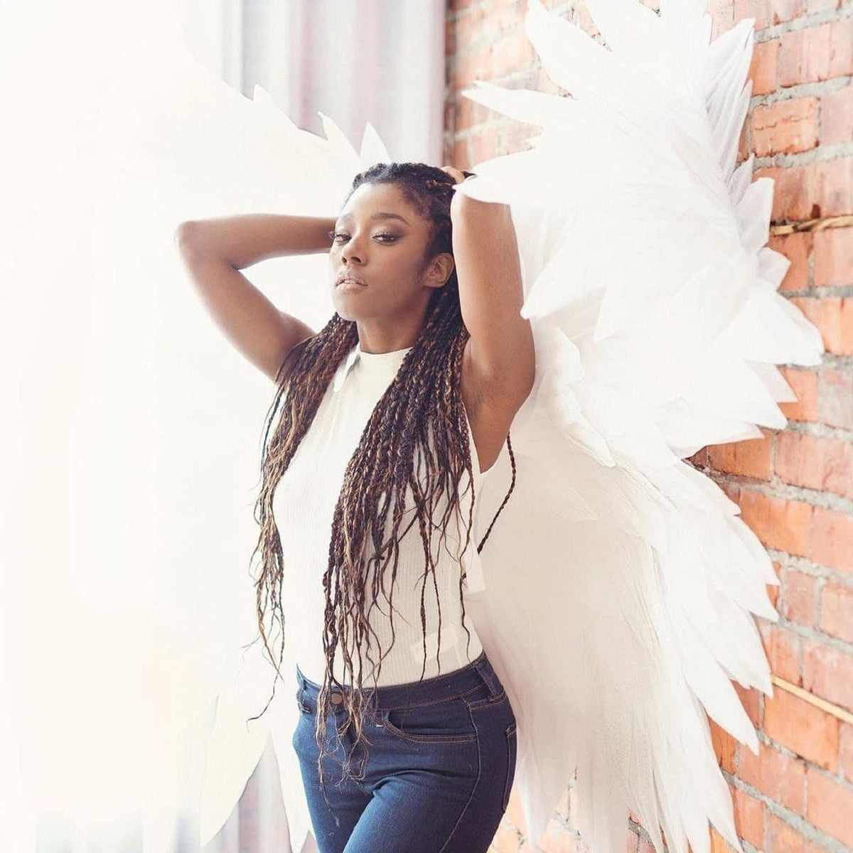 May include: A woman with long brown braids wears a white sleeveless top and blue jeans. She is standing in front of a brick wall with large white angel wings behind her.