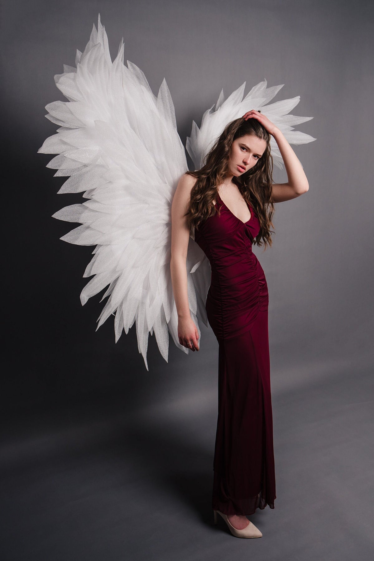 May include: A woman wearing a long, burgundy dress with a pair of white angel wings behind her. She is looking at the camera with a serious expression.