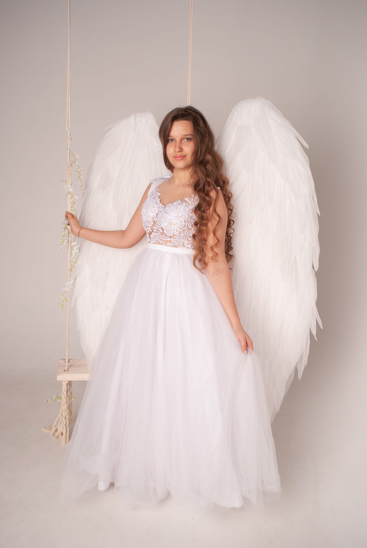May include: A young woman wearing a white lace dress and large white angel wings. She is standing in front of a white background.