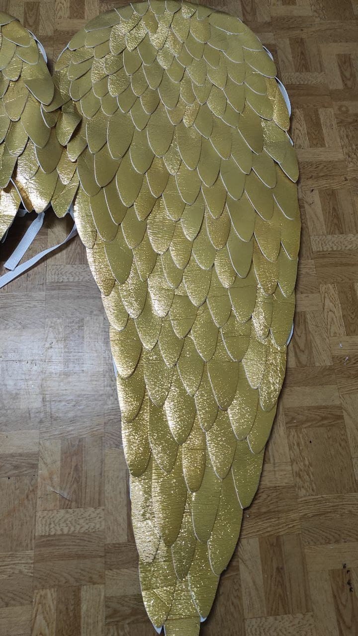 May include: A pair of large decorative wings featuring a layered design of metallic gold, feather-shaped cutouts. The wings are constructed with a white base layer and include white elastic straps attached to the upper section for securing the item.