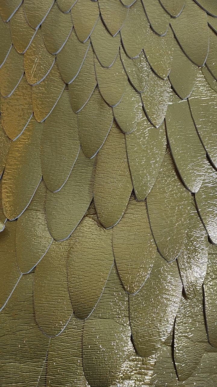 May include: A pattern of overlapping, teardrop-shaped scales in a metallic gold finish. Each individual scale features a fine, horizontal ribbed texture and a thin white edge, creating a layered, repetitive design across the entire surface.