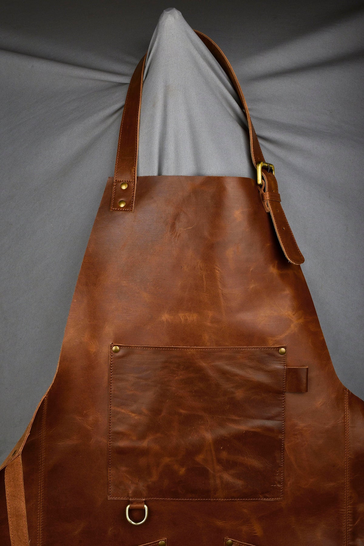 May include: A brown leather apron with a large pocket and adjustable straps. The apron has brass hardware and a distressed look.
