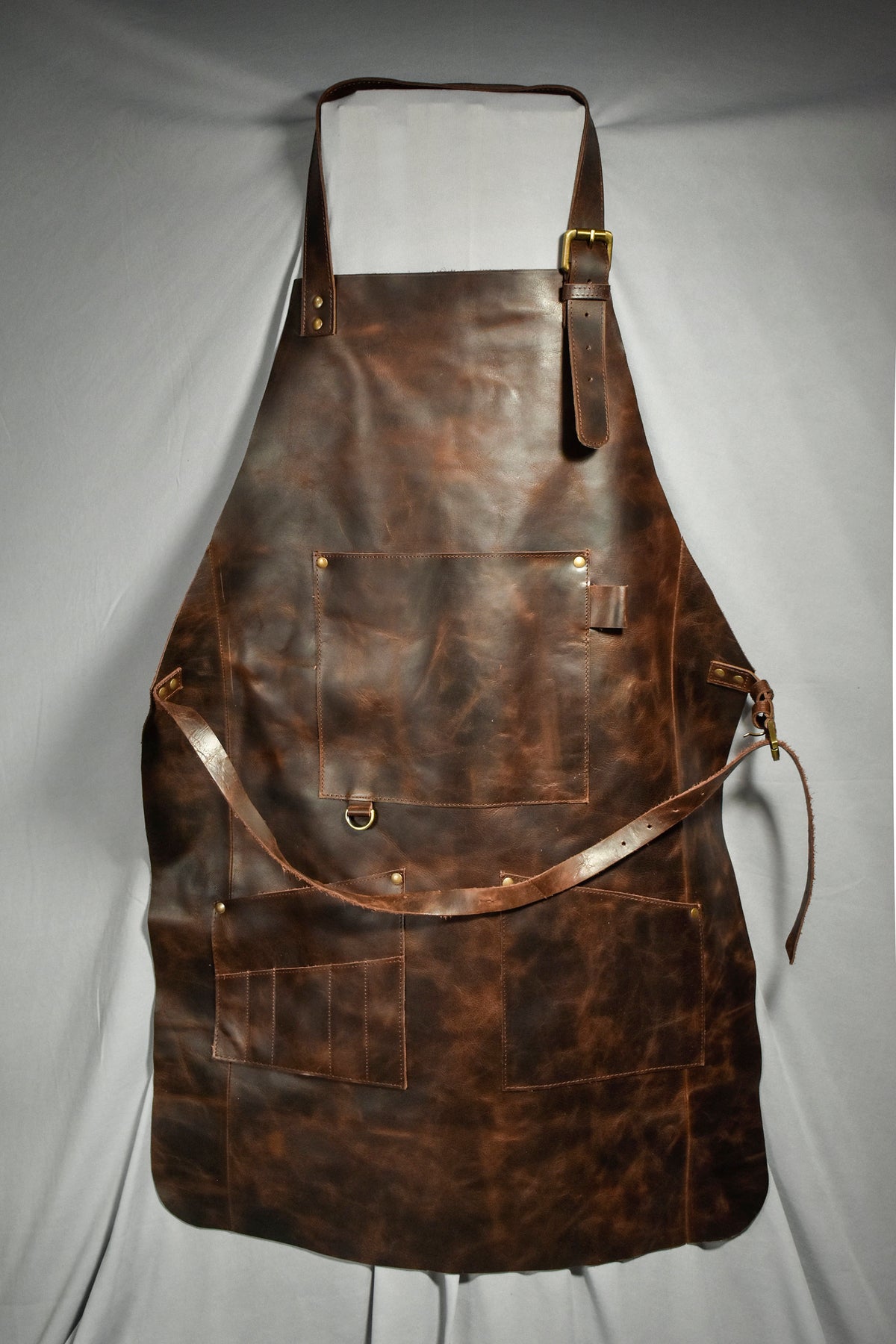 May include: A brown leather apron with multiple pockets and adjustable straps. The apron features a chest strap with a buckle and a waist strap. The leather has a distressed look with visible grain and variations in color. The apron is designed for protection and utility.