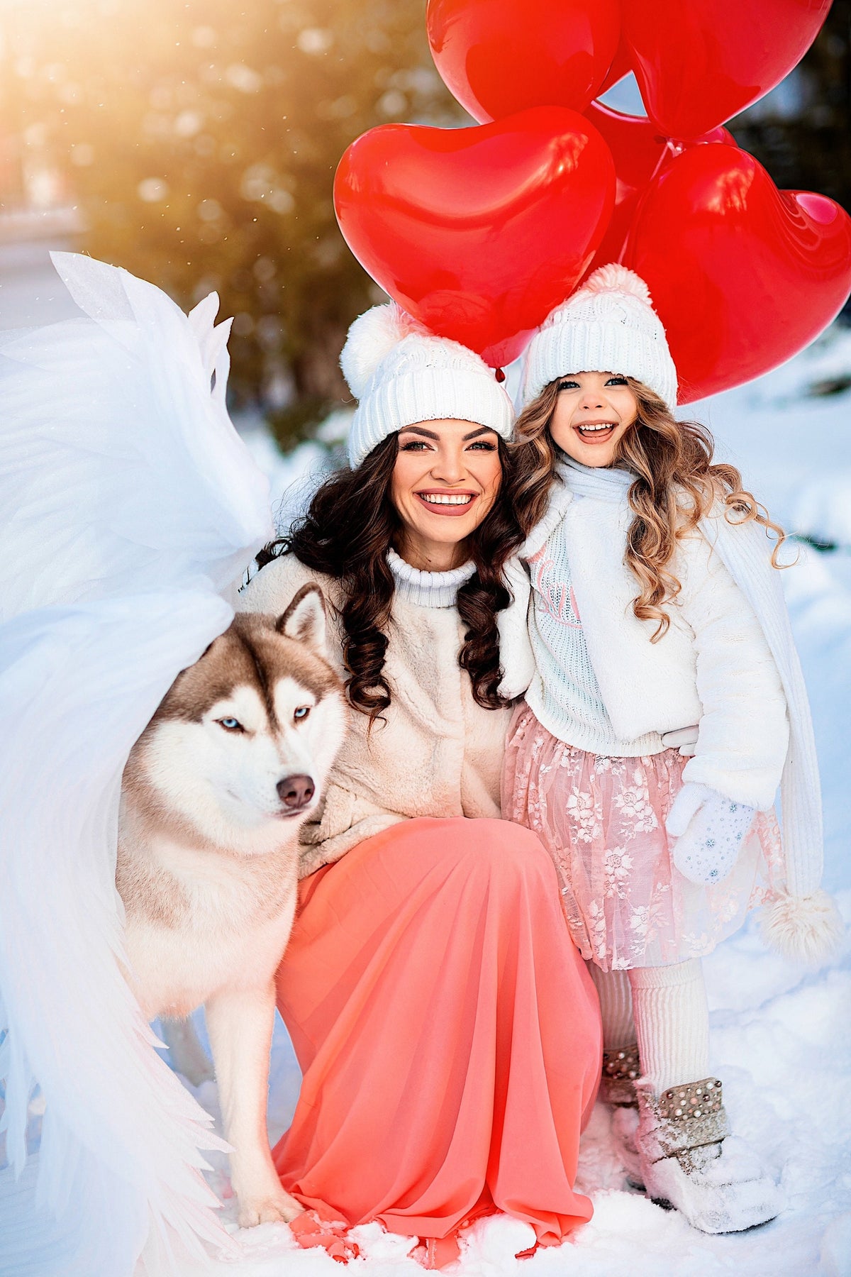 May include: A woman and a young girl wearing white winter hats and coats pose with a Siberian husky dog in a snowy forest. They are holding red heart-shaped balloons.