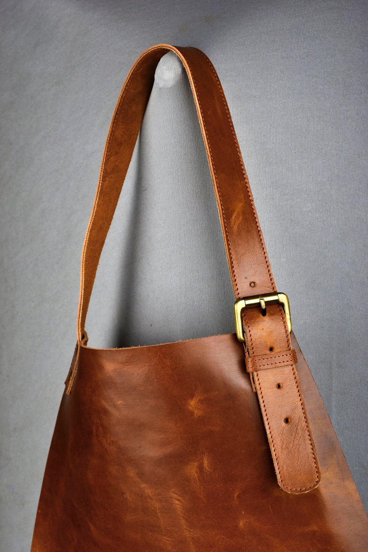 May include: A brown leather apron with a long, adjustable strap. The apron features a brass buckle and a simple, functional design. The leather has a natural, textured appearance, suggesting durability and quality. The apron is designed for use in a kitchen or workshop.