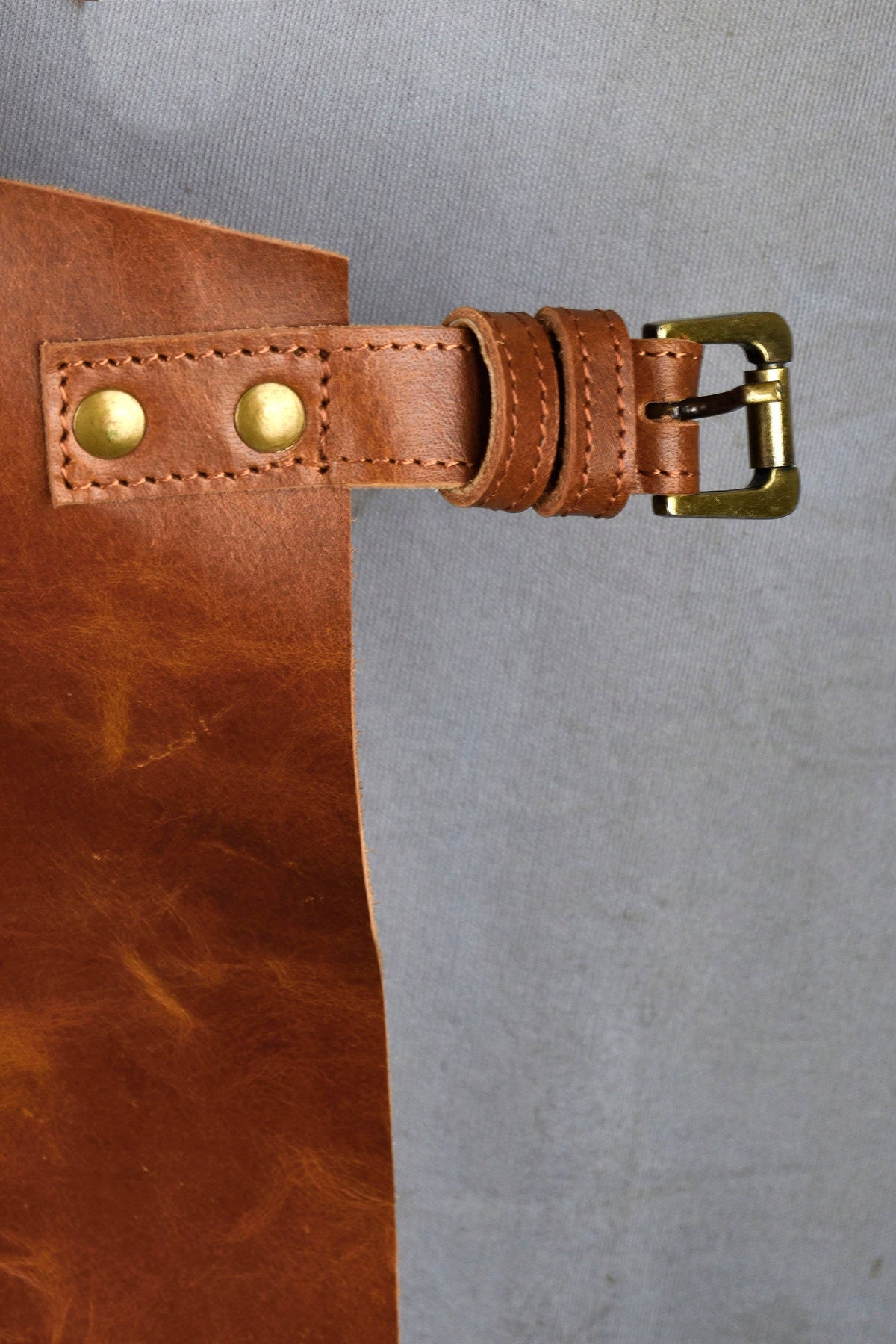 May include: A brown leather apron with a brown leather belt and a brass buckle. The apron has two brass rivets on the belt.