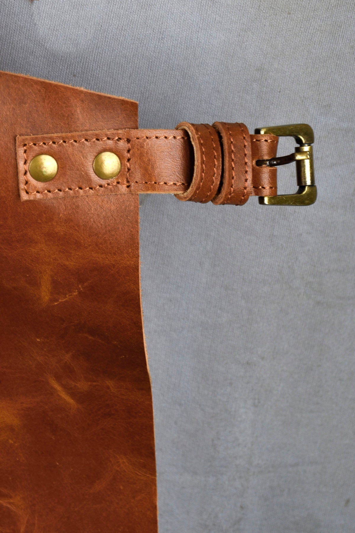 May include: Close-up of a brown leather apron detail. The apron features a leather strap with a brass buckle and two brass rivets. The leather has a textured appearance and is stitched with a contrasting thread. The apron is designed for use in the kitchen or workshop.