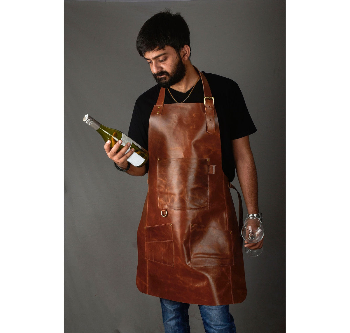 May include: A brown leather apron with adjustable straps and a large pocket. The apron is worn by a person holding a bottle of wine and a glass.