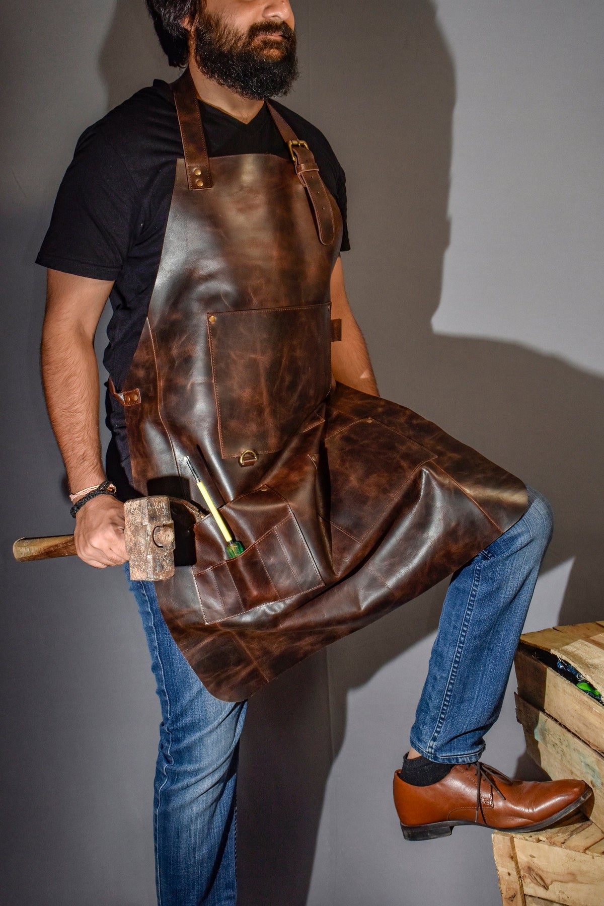 May include: A brown leather apron with multiple pockets and a hammer in the front pocket. The apron is worn by a person wearing a black t-shirt and blue jeans.