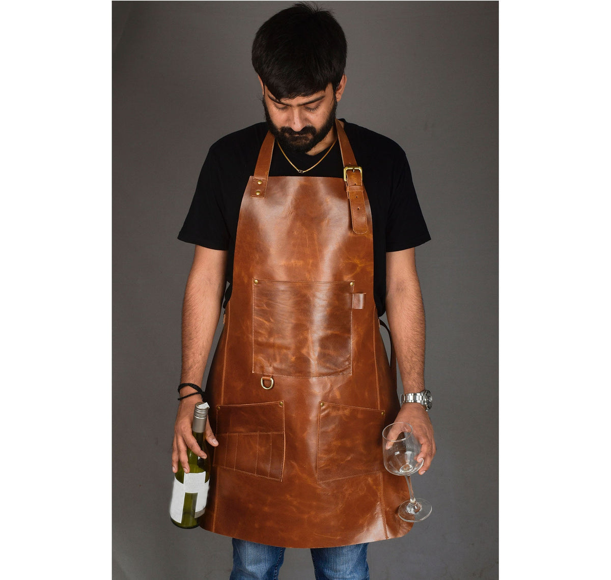 May include: A brown leather apron with multiple pockets and brass hardware. The apron has a neck strap and waist ties. The apron is worn by a person holding a wine bottle and a wine glass. The apron is designed for use in a kitchen or bar setting.