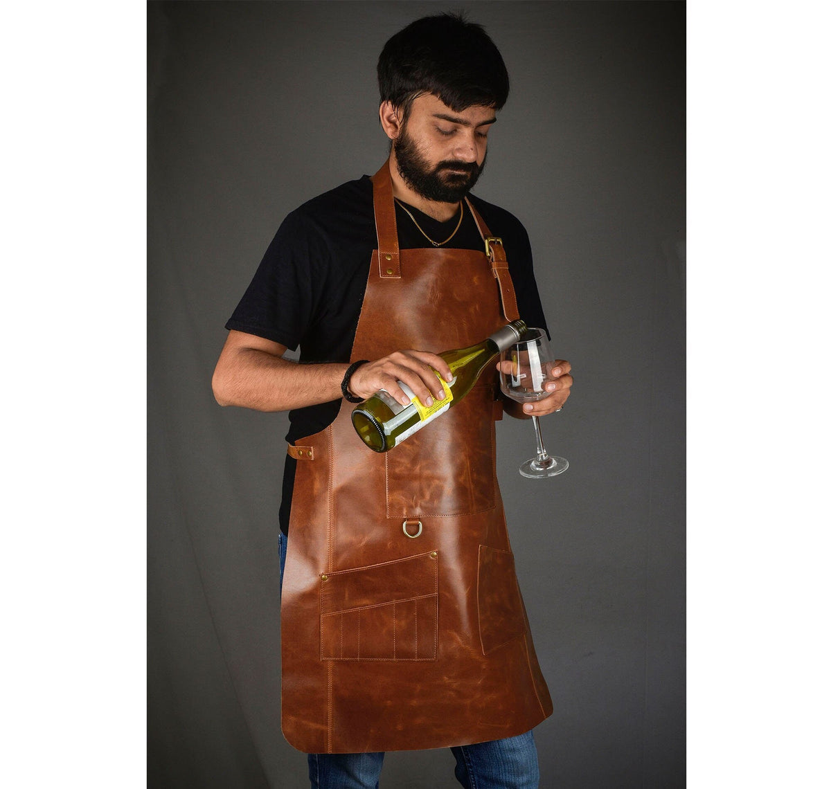 May include: A brown leather apron with multiple pockets and brass hardware. The apron is being worn while pouring wine into a clear wine glass. The apron has a neck strap and waist straps.