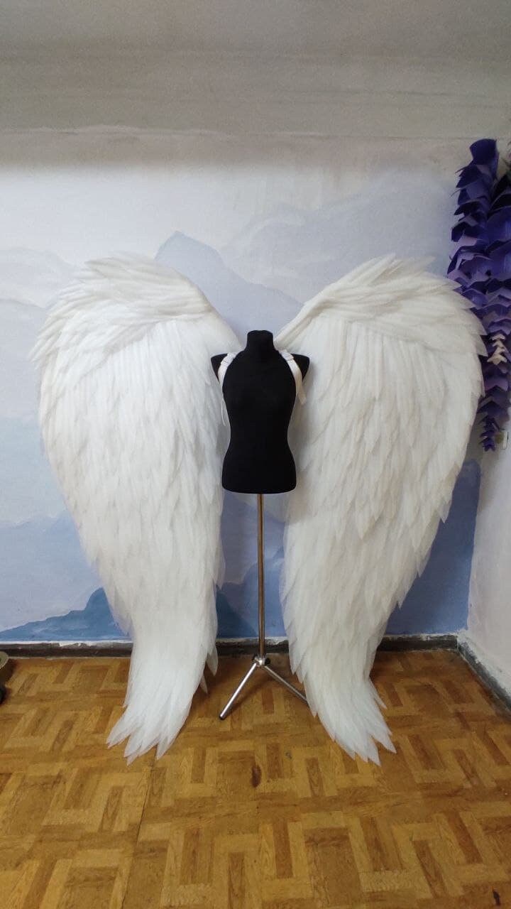 May include: A pair of large, white angel wings made of feathers. The wings are attached to a black mannequin and are displayed against a light blue and white background.