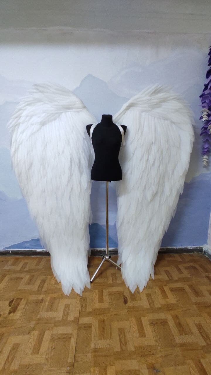 May include: A pair of large, white angel wings made of feathers. The wings are attached to a black mannequin standing on a silver stand. The wings are displayed against a light blue wall with a white mountain scene painted on it.