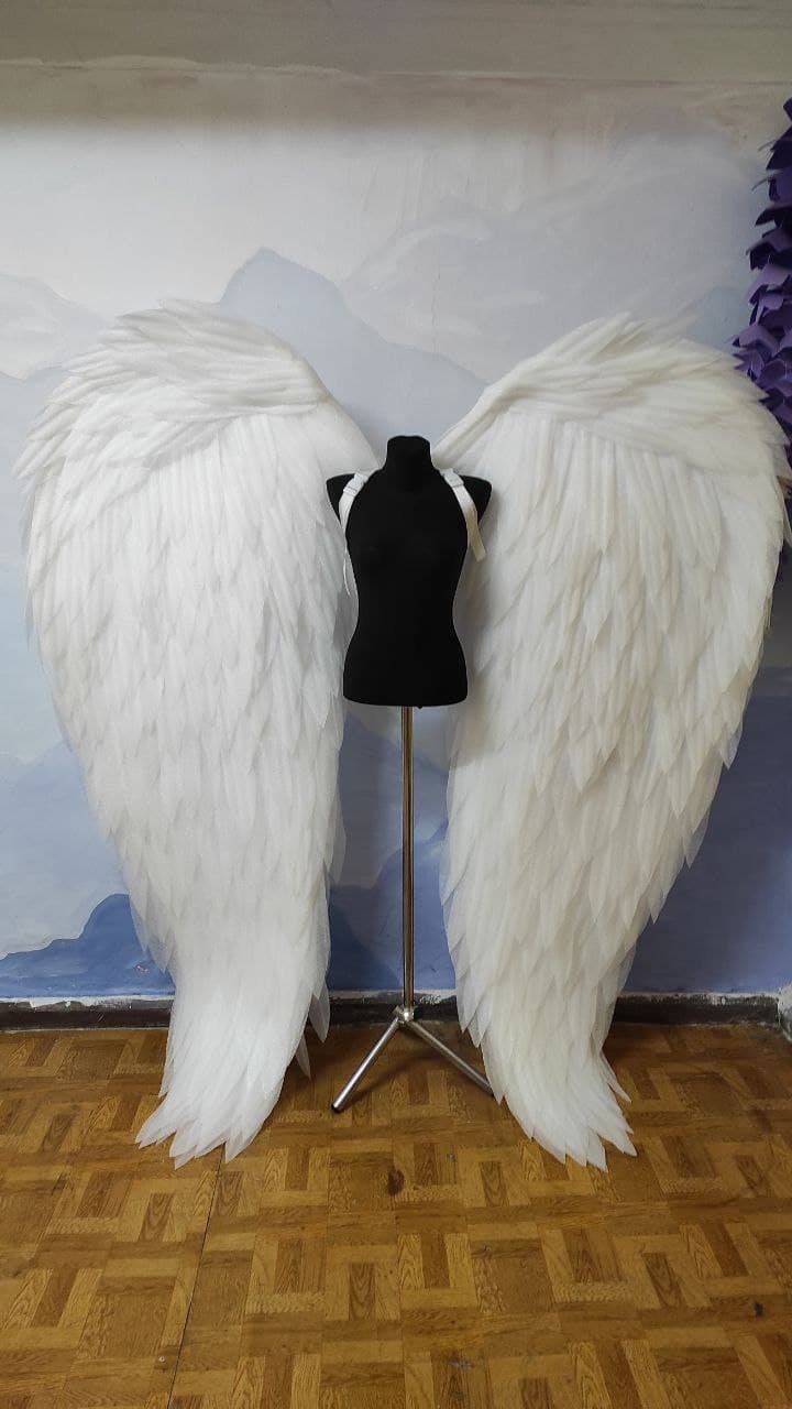 May include: A pair of large, white angel wings made of feathers. The wings are attached to a black mannequin torso on a silver stand. The wings are displayed against a light blue and white background.