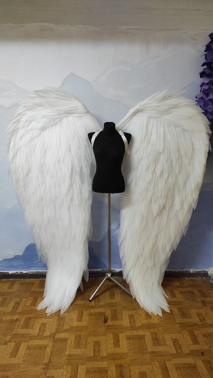 May include: A pair of large, white angel wings made of feathers, displayed on a mannequin. The wings are attached to a stand and are positioned against a light blue and white background.