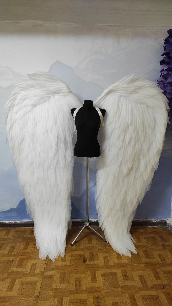 May include: A pair of large white angel wings with a feathered texture, displayed on a black mannequin stand against a white wall.