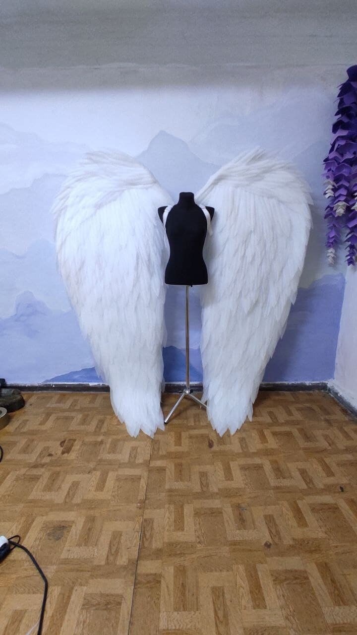 May include: A pair of large, white angel wings made of feathers, displayed on a black mannequin stand against a blue and white background.